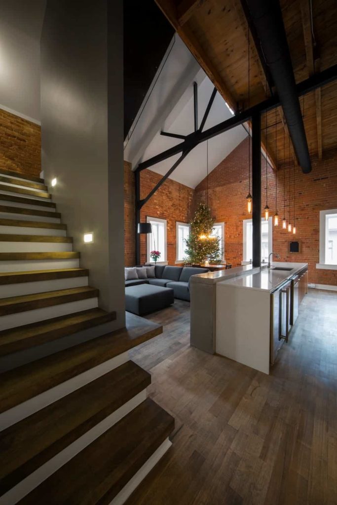 This Industrial-style living room is within a great room that also houses both the kitchen and dining area under its large cathedral ceiling with exposed metal beams and its tall red brick walls with contrasting white windows that bring in natural lights L-shaped gray sectional sofa of the living room with a matching cushioned ottoman.