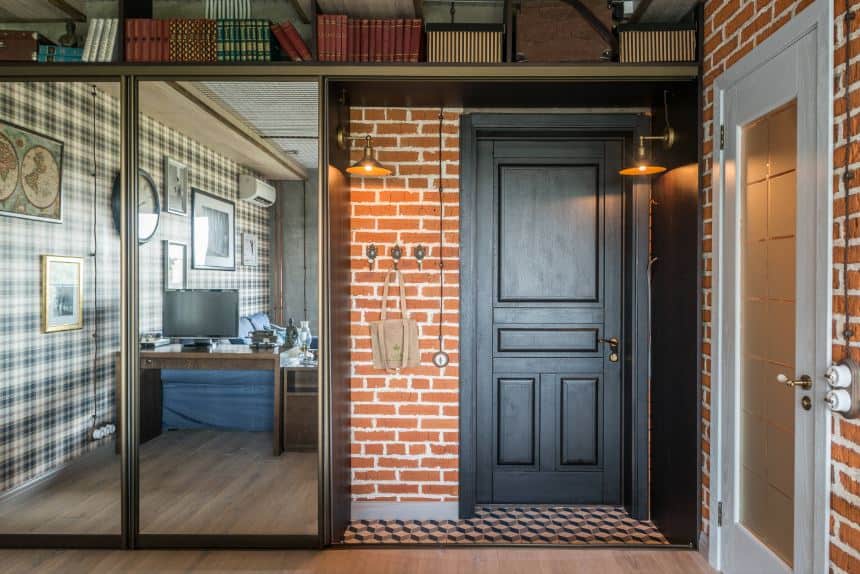 This charming and homey industrial-style foyer has wall-mounted lamps flanking the dark wooden door complemented by red brick walls with the vertical space maximized through book shelves.