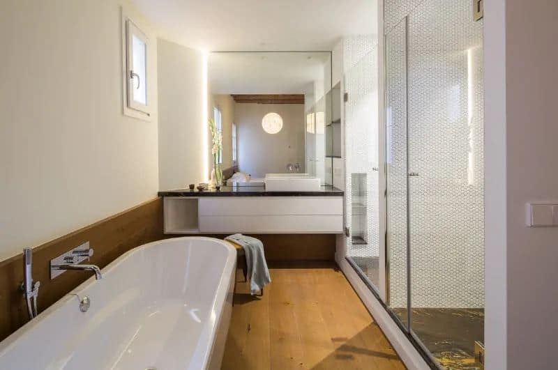 This simple bathroom has a long elliptical freestanding bathtub that stands out against the brown hue of the flooring and backsplash. Across from this are two glass-enclosed shower areas with small tiles on its walls and black marble on its flooring.
