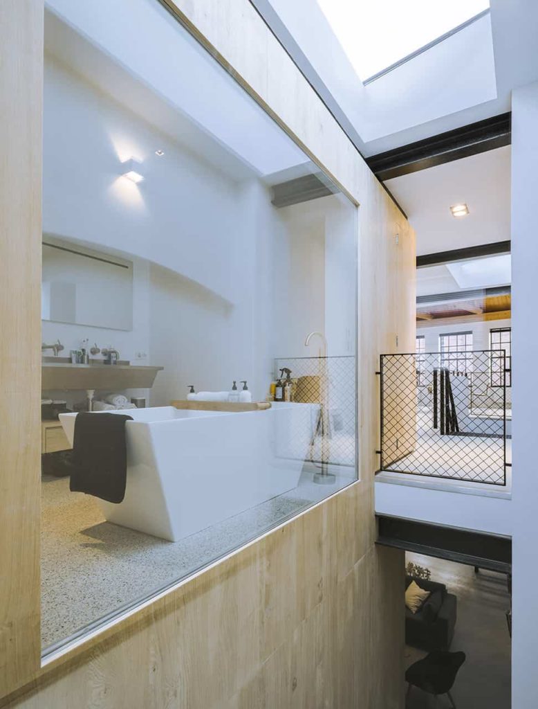 The simple bathroom can be clearly seen from the staircase through its glass wall. It has a white freestanding bathtub across from the gray floating two-sink vanity with exposed pipes underneath that matches the faucets.