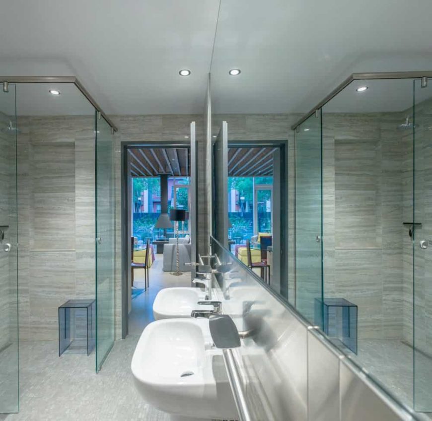 This bathroom has two floating white sinks with stainless steel faucets complementing the large vanity mirror embedded into the wall. This works well with the glass enclosure of the shower area that has gray flooring tiles.