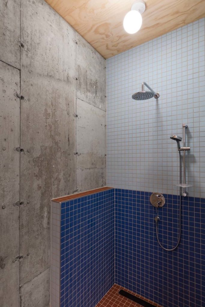 This bathroom has a shower area with small blue tiles on its walls that stand out against the gray concrete walls and bare wooden ceiling that supports a large white flush-mount light that illuminates the stainless steel fixtures of the shower area.