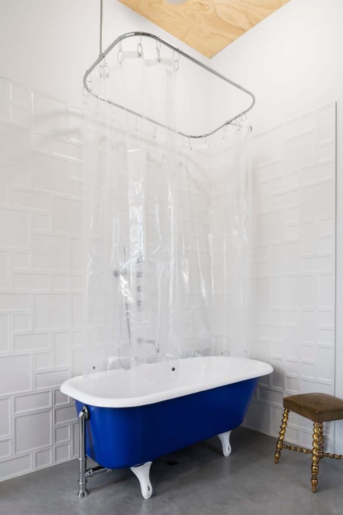 This bathroom has a blue and white freestanding bathtub in a corner of white patterned walls and gray concrete flooring. This bathtub also acts as a shower area with a see-through plastic shower curtain under a bare wooden ceiling.
