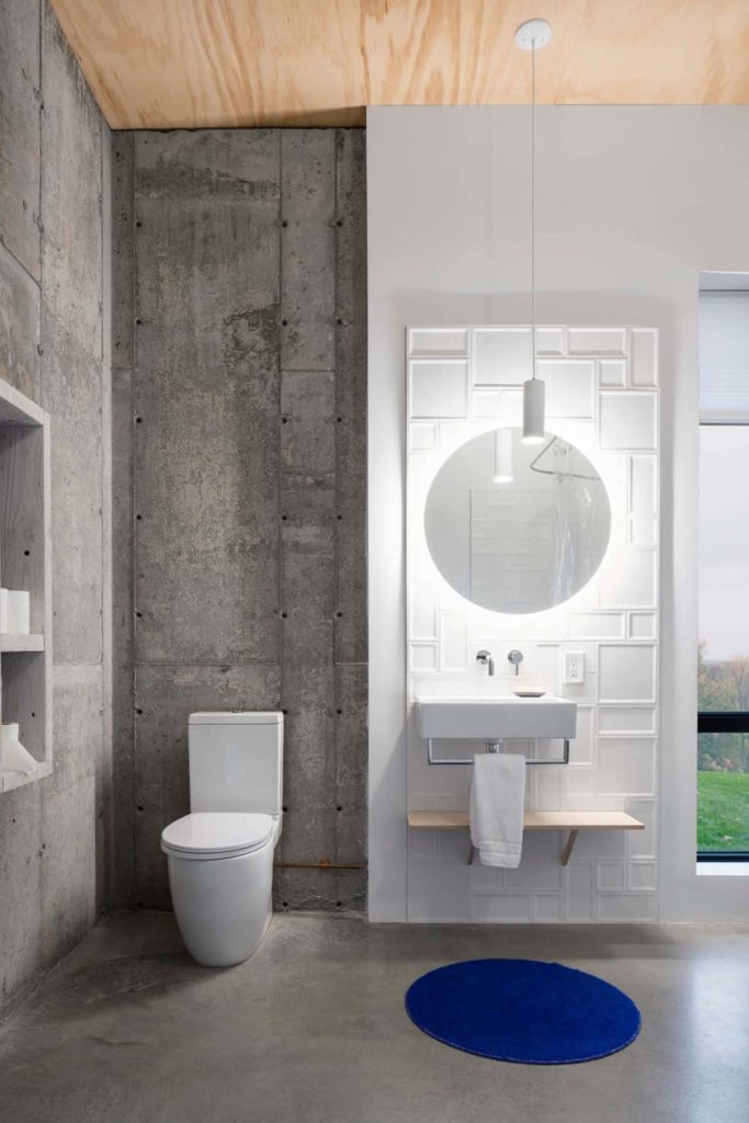 This simple bathroom has a bare wooden ceiling paired with gray concrete walls and flooring that contrasts the white modern toilet and the sink that is against a modern wall panel that is white, patterned and lit with white lights.