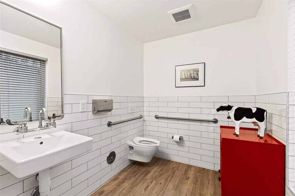 This simple bathroom has a hardwood flooring that complements the white wall tiles arranged in a brick wall patter that houses the floating porcelain toilet and sink with exposed pipes underneath. This makes the red drawer stand out adorned with a cow figurine.