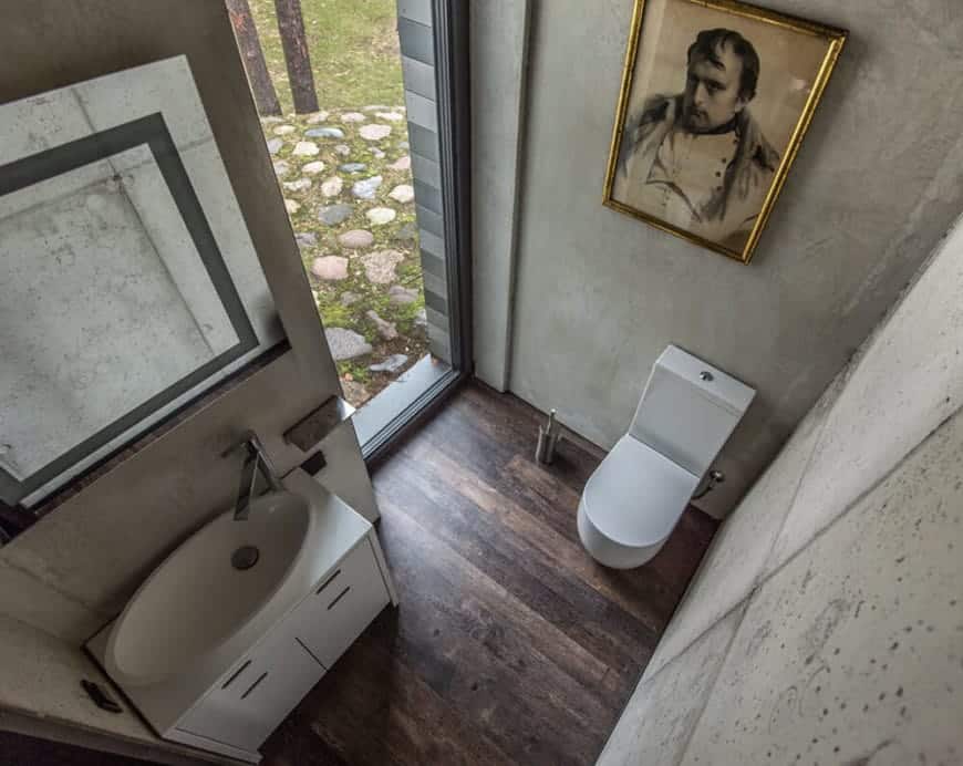 The white modern toilet and floating white vanity stands out against the gray concrete walls accented with a classic painting above the toilet and a wall-mounted vanity mirror above the white sink. The dark hardwood flooring is illuminated by the tall and narrow glass window beside the toilet.