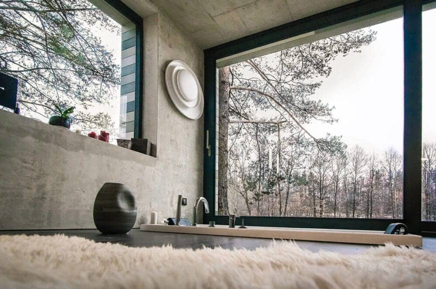 This industrial-style primary bathroom has a bathtub embedded into the black flooring that is complemented by the gray concrete walls and ceiling. These are all illuminated by the natural lights coming in from the wide glass windows.