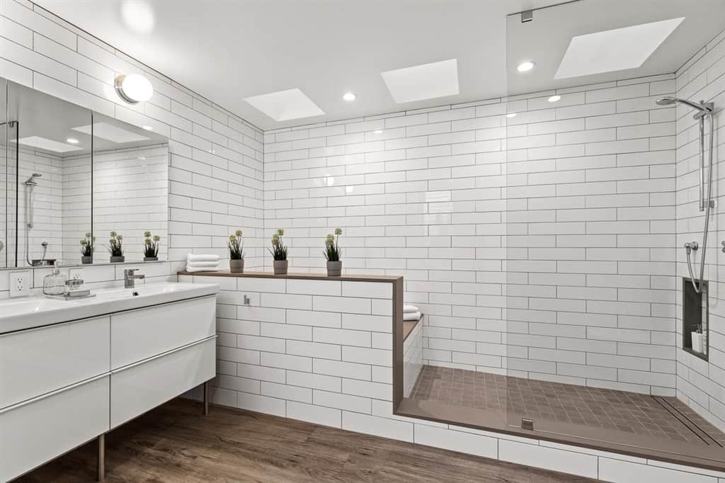 This bathroom has enough space for a bathtub and a shower area on the far wall that is made up of white tiles arranged in a brick wall pattern complemented by the dark wood hue of the flooring tiles of the shower area matching with the hardwood flooring of the vanity area.