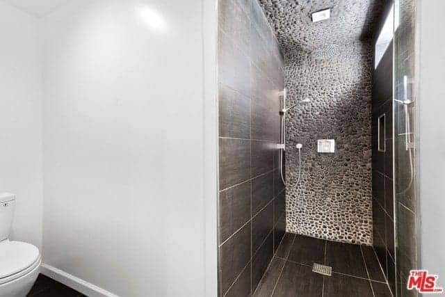 The white porcelain toilet is placed in a corner of white walls. Adjacent to this is the glass door of the shower area that has contrasting dark tiles on its floor and walls except the far wall and ceiling that is made of small stones embedded in concrete.