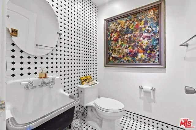 A nice dash of color is provided by the colorful abstract painting mounted on the white wall beside the white toilet that stands out against the patterned black and white hues of the flooring tiles that extend to the wall behind the industrial-style sink and toilet.
