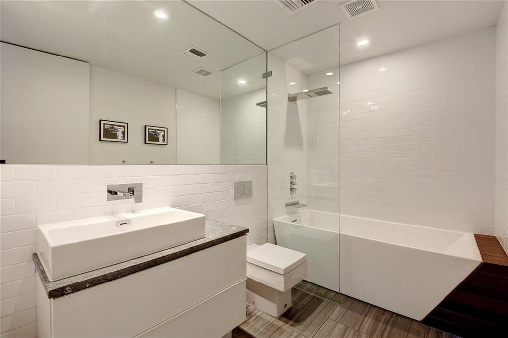 The porcelain white sink with its own built-in countertop matches with the white porcelain toilet beside it that stands out against the dark industrial-style tiles of the flooring that extends to the small glass-enclosed shower area.
