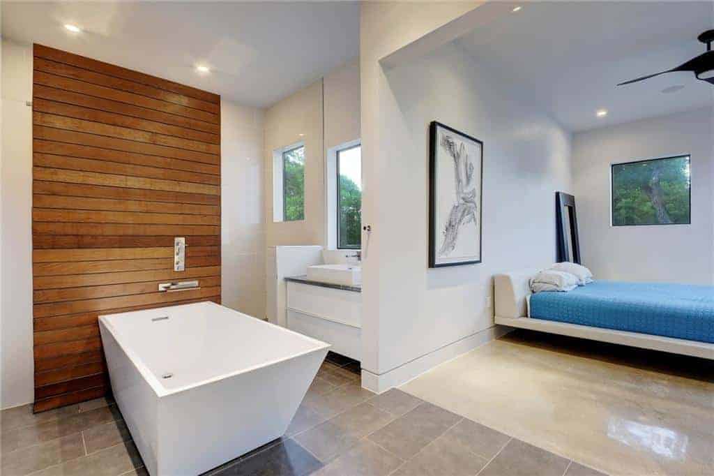 The rectangular freestanding white bathtub that stands out against the gray industrial-style flooring and its wall panel made of wooden planks embedded into the white wall. This setup is illuminated by the window above the modern white vanity.