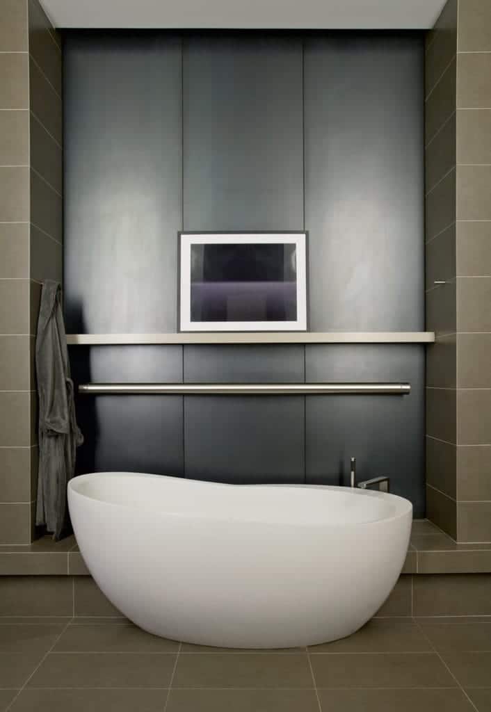 The white freestanding bathtub is paired with a stainless steel faucet matching the stainless steel rod that stands out against the black wall panel that is surrounded by the gray tiles of the flooring and the walls.