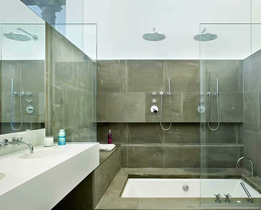 The white floating sink of this industrial-style bathroom matches with the white bathtub that is embedded into gray concrete that extends to the walls of the shower area that has a half glass wall and overhead shower head.