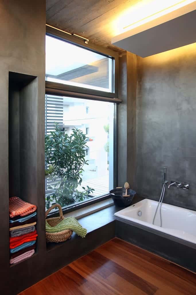 This simple bathroom has gray concrete walls that houses the white bathtub and built-in shelves. This matches with the gray concrete ceiling that has a white wooden panel over the bathtub that has lighting.
