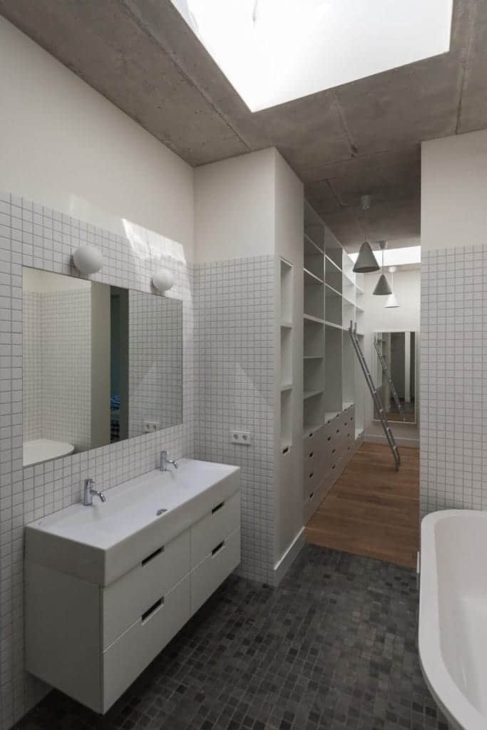 This simple bathroom has small tiles on its walls and flooring. The flooring has various shades of gray while the walls have only white tiles. These are complemented by the gray concrete ceiling that has a skylight over the white bathtub and vanity.