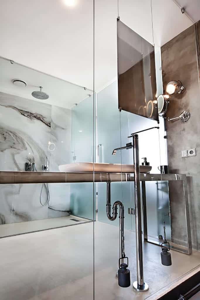 This bathroom is seen from the outside through a glass wall. It has a gray concrete flooring that complements the stainless steel vanity and the pipes of the white sink as well as the fixtures of the glass-enclosed shower area across.