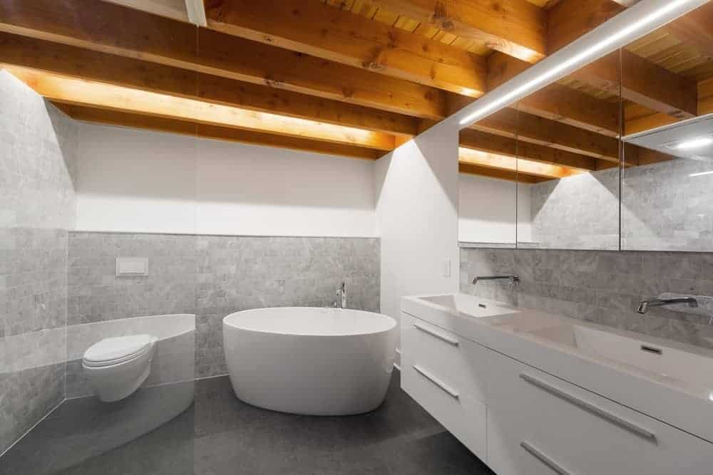 This industrial-style bathroom has a wooden ceiling with exposed wooden beams that contrast the white walls. These walls are dominated by the gray backsplash tiles that complement the white toilet, freestanding bathtub and the floating modern two-sink vanity.