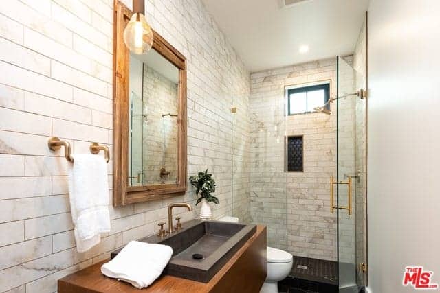 This bathroom has beautiful white marble tiles that are arranged in a brick wall pattern. These tiles extend from the walls of the vanity area to the shower area that has a glass door. The vanity area is adorned with a wood framed mirror and brass fixtures.