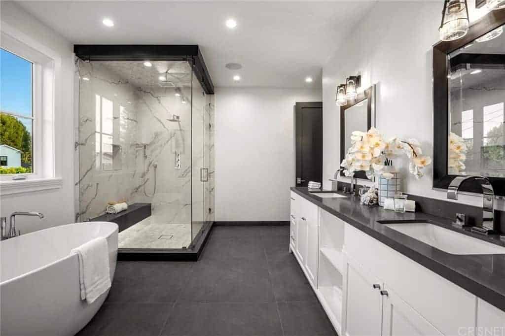 The dark gray countertop of this primary bathroom matches with the dark gray tiles of the flooring. This is then contrasted by the white freestanding bathtub and the white wooden vanity that has shaker cabinets and drawers topped with vanity mirrors.