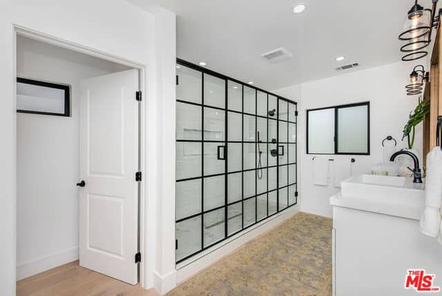 This primary bathroom has a wide shower area enough for two within its glass wall that has black frames that matches with the window and the fixtures of the shower area and vanity area. This has a white sink topped with a wood-framed mirror.