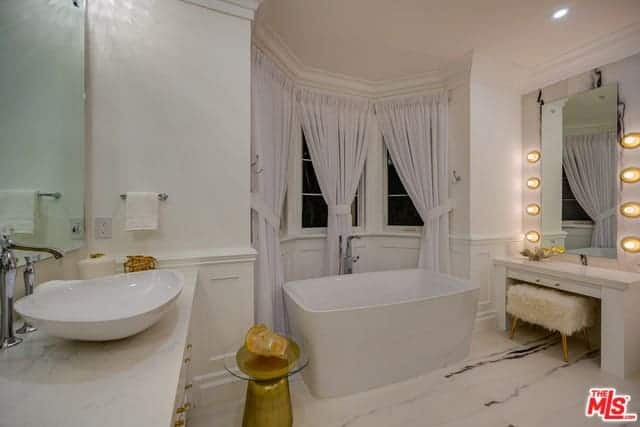 The charming nook alcove of curtained windows and white wainscoting is a nice background for the freestanding bathtub that blends with the white marble flooring. Beside this is the white vanity area with a large mirror bordered with yellow spherical lights.