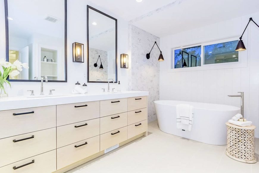 The bright white freestanding bathtub, walls and ceiling of this Farmhouse-style bathroom is complemented by the beige wooden vanity that matches with the flooring and the stool beside the bathtub. These are augmented by the dark wall-mounted lamps.