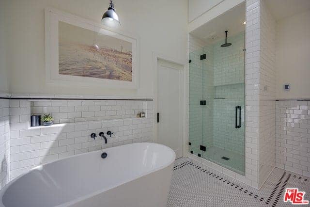 There is a beautiful colorful painting mounted on the beige upper wall by the freestanding white bathtub with a white-tiled backsplash contrasted by the black faucet. This matches with the fixtures of the shower area that has a glass door.