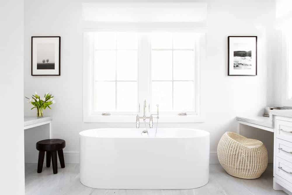 The bright French windows bring in an abundance of natural lights that lighten up the white walls accented with framed artworks. It also lightens up the white vanity and freestanding bathtub adorned with a rustic woven basket chair beside it.