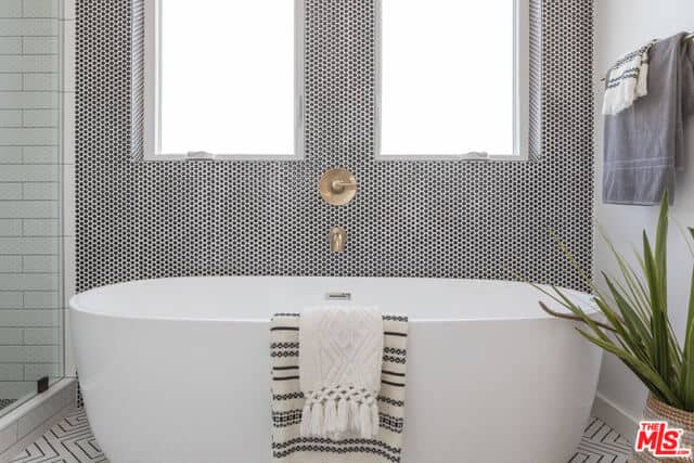 The white freestanding bathtub stands out against the wall that has a complex pattern to it contrasted by the golden faucet in between of the two windows that bring in an abundance of natural lighting to the white patterned flooring and potted plant.