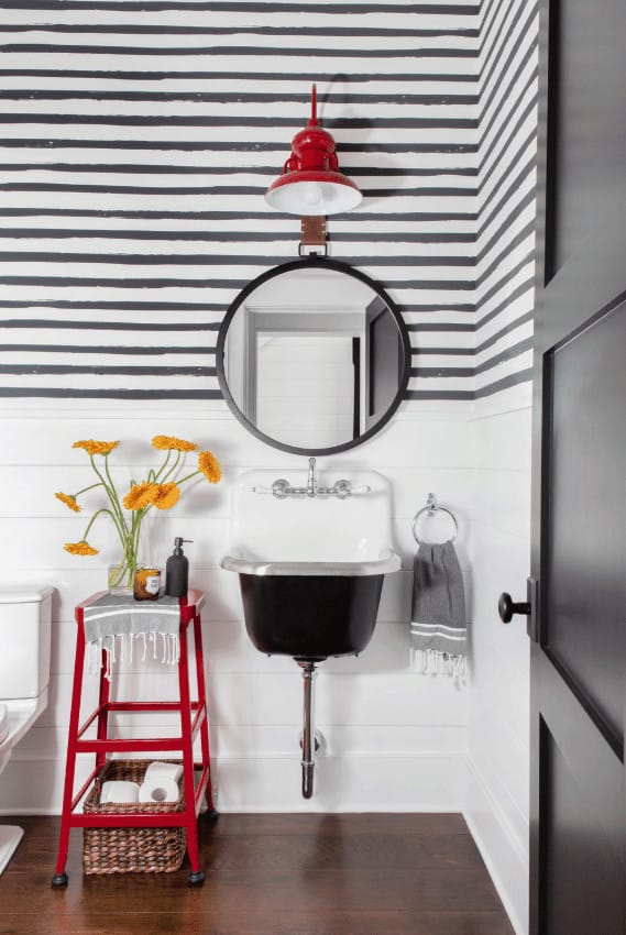 There is a red metal stool that stands out against the white wooden wainscoting that has a wooden plank finish. This stool matches with the red Farmhouse-style wall lamp above the round mirror mounted on the upper walls that have a striped wallpaper finish.