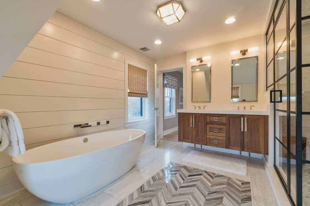 The white freestanding bathtub is against a wall with a white wooden plank finish warmed by the yellow lights of the wall-mounted lamps above the vanity mirrors above the wooden vanity that has shaker cabinets and drawers that stand out against the light hue of the flooring.