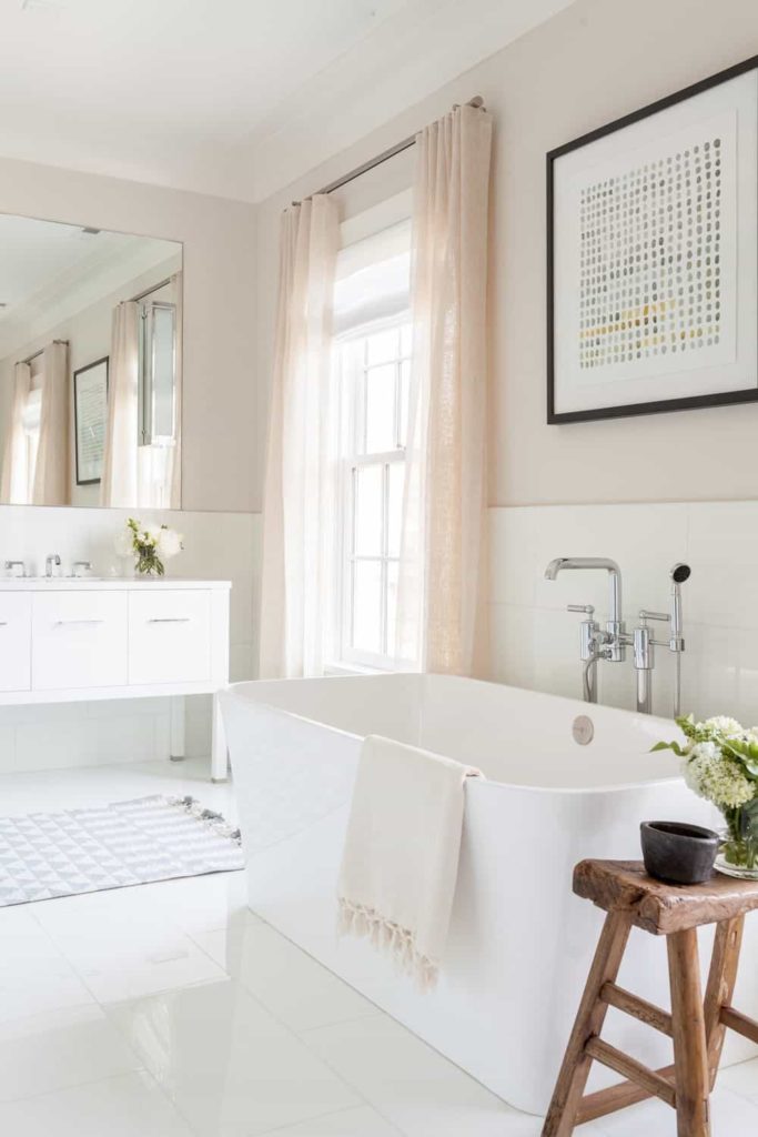 The bright natural lights that are coming in from the pink curtained window brightens up the white wooden cabinets of the vanity, the white tiles of the flooring and the white freestanding bathtub adorned with a small wooden stool beside it that serves as a table.