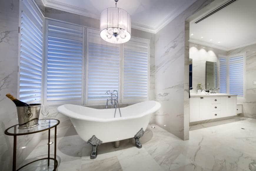 The elegant freestanding bathtub has silvery legs with intricate carvings on it. This is placed in an alcove of white marble walls and flooring surrounded by white shuttered windows topped with a small white chandelier with a round hood on it.
