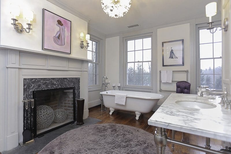 This is an elegant and warm Farmhouse-style primary bath that has a white freestanding bathtub that stands out against the dark hardwood flooring beside the fireplace with a light gray mantle topped with a colorful painting across from the vanity area.