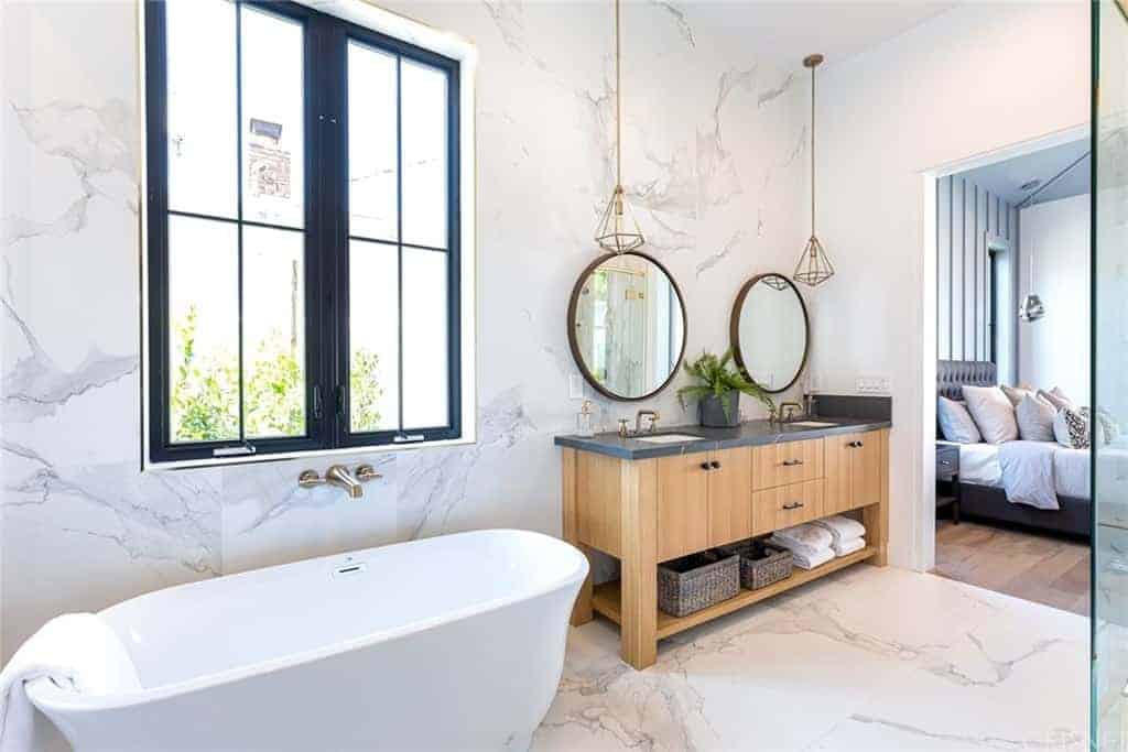 The white marble flooring tiles extends to the walls and backsplash of the white freestanding bathtub and the two-sink vanity that has cabinets, drawers and a shelf at the bottom for the towels and baskets. This is paired with round mirrors and pendant lights.