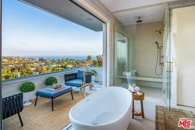 The wall beside the freestanding bathtub opens up to a bright veranda that has a sitting area of armchairs and coffee table with blue cushions. This brings in an abundance of natural lighting to the glass-enclosed shower area by the head of the bathtub.