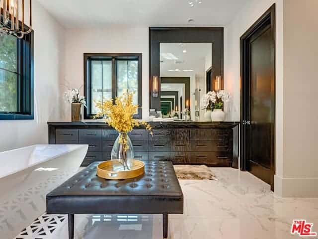 There is a dark brown leather tufted bench beside the white freestanding bathtub that matches with the dark wooden hues of the vanity drawers as well as the door next to it and the frame of the large wall-mounted vanity mirror with wall-mounted lamps on its frame.