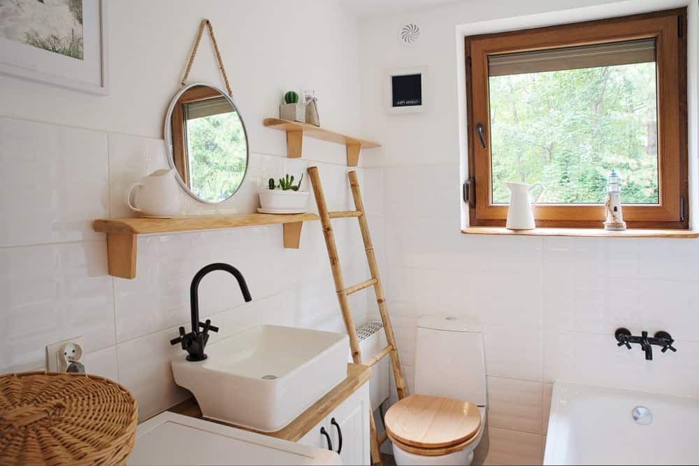 This is a small and simple Farmhouse-style bathroom that has a white porcelain toilet that has a wooden cover matching with the wooden hues of the ladder beside it, vanity countertop, shelves and the frames of the window above the head of the bathtub.