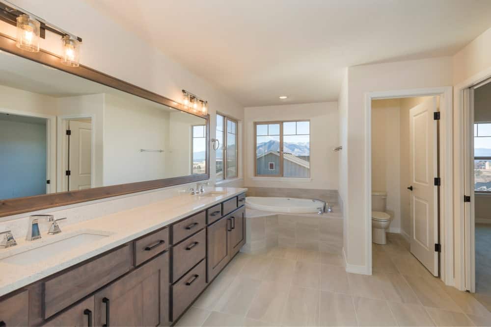 The large two-sink vanity is the highlight of this Farmhouse-style primary bathroom with a corner bathtub by the windows beside the toilet area that has its own white wooden door. This matches with the light hues of the flooring tiles.