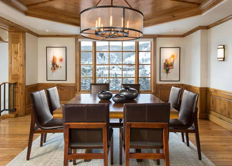 The charming brown leather cushioned chairs are a perfect complement for the wooden dining table that matches with the wooden wainscoting of the white walls. This aesthetic is mirrored by the white ceiling that has a wooden central tray.