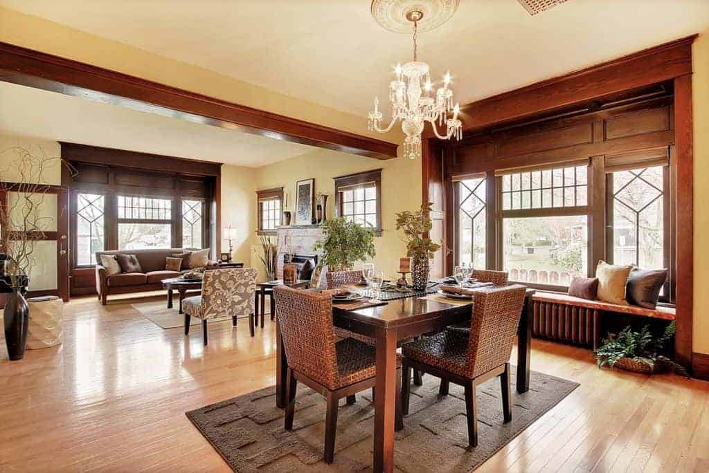 The wooden rectangular table is paired with woven wicker chairs of the same hue over a dark gray area rug of the light hardwood flooring. The table matches with the large wooden wall that houses the windows and a built-in cushioned bench for a sitting nook.
