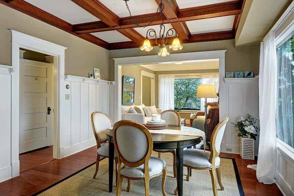 The hardwood flooring that is topped with a rustic woven area rug matches with the exposed wooden beams of the white ceiling. This is reflected by the white wainscoting contrasted by the green upper walls of this medium-sized dining room.