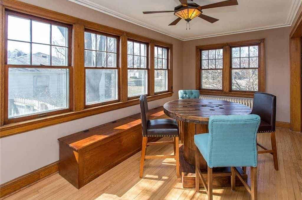The highlight of this medium-sized dining room is the rustic round wooden table that is a repurposed cable reel. This matches the warm homey quality of the light hardwood flooring, long bench with storage and the frames of the windows that bring in natural lights.