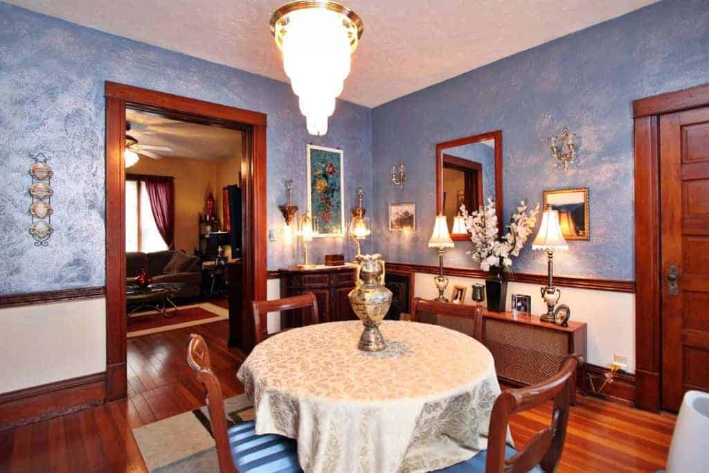 This medium-sized dining room balances on the thin line separating elegance and drama with its light blue textured walls that serve as a nice background for the various decorations, artworks and wall-mounted lamps. These are paired with a simple round table with a white tablecloth and wooden chairs.