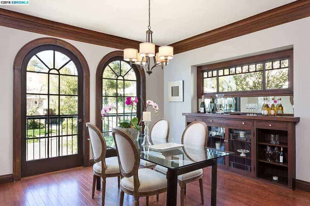 The hardwood flooring of this medium-sized dining room matches with the frames of the arched glass doors that bring in an abundance of natural lights to the glass-top dining table paired with white cushioned oval back chairs that stand out against the flooring and dining room cabinet embedded into the white wall.