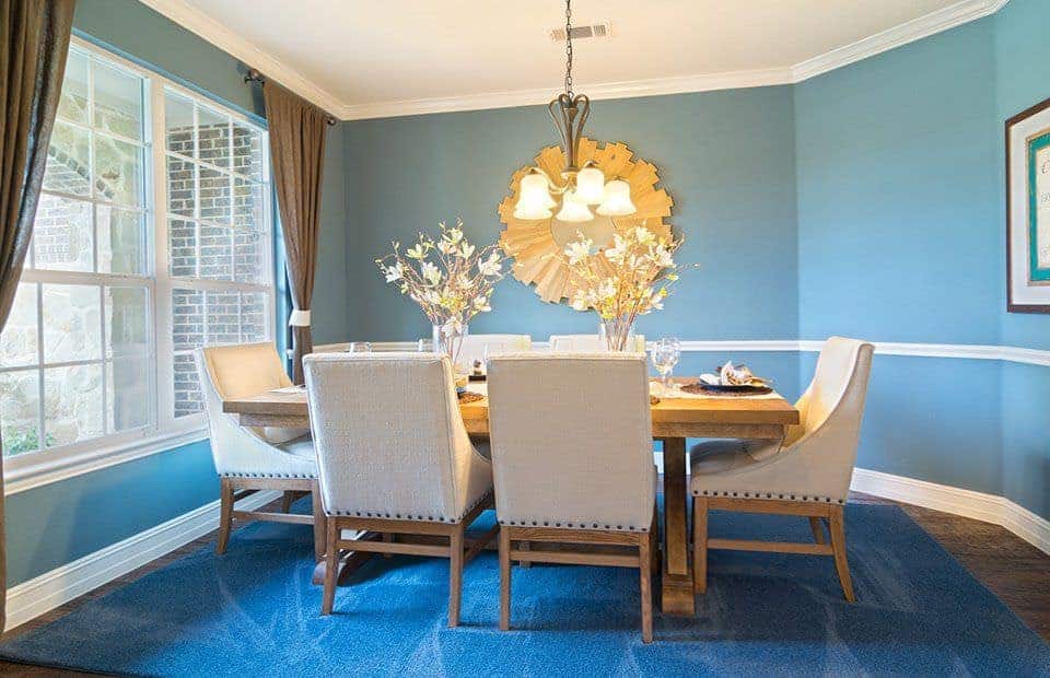 This medium-sized dining room has a rectangular wooden table in the middle of the blue area rug encircled by light gray cushioned chairs. This is given a nice background of light blue walls and white ceiling brightened by the chandelier and the French windows.