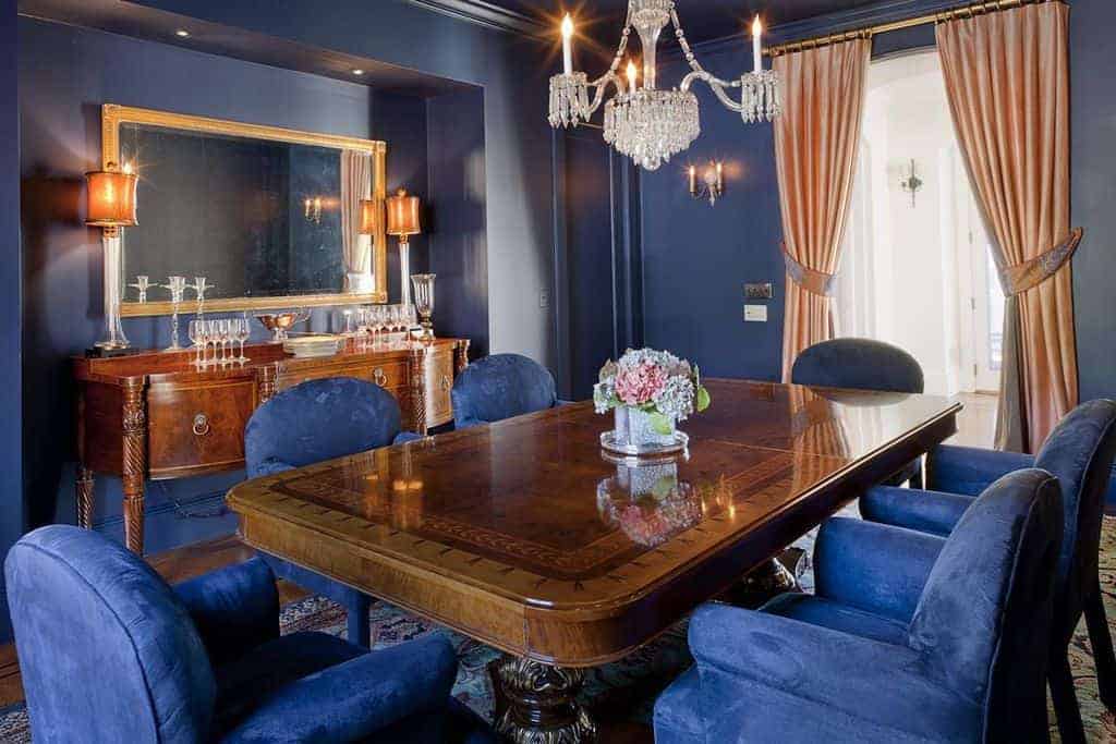 The blue walls of this medium-sized dining room blend with the blue velvet cushions of the armchairs that encircle the wooden dining table that matches the console table. The dining table is adorned with a simple crystal chandelier hanging from the blue ceiling.