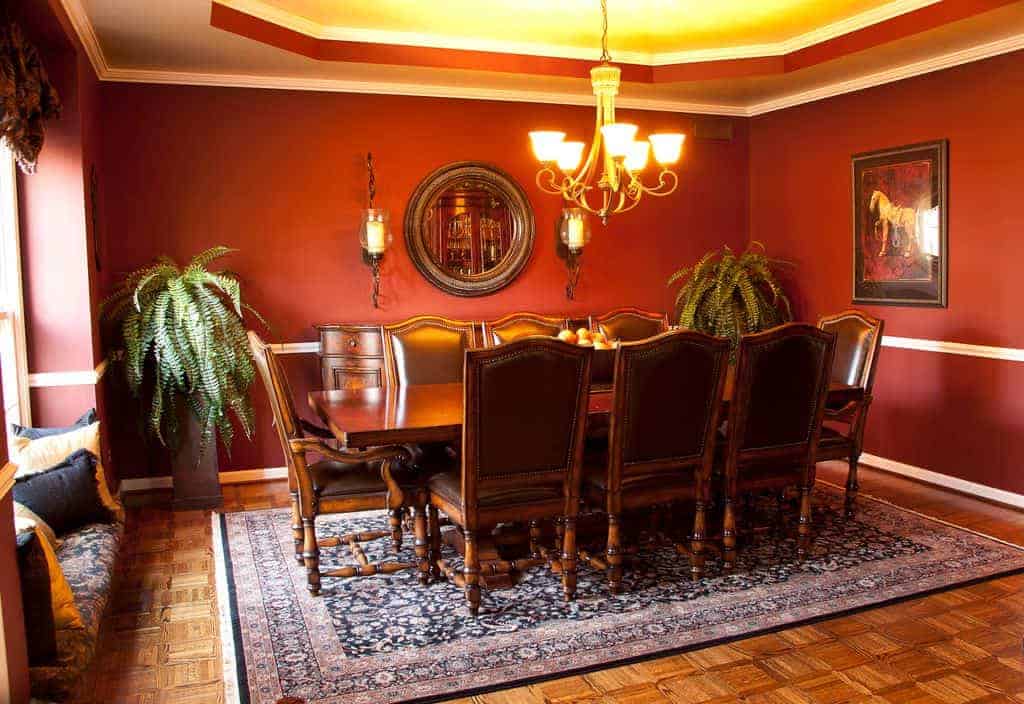The antique-style rectangular wooden dining table is paired with brown leather cushioned chairs. These are a match with the dining room cabinet and the round wall-mounted mirror above it brightened by the warm yellow lights of the chandelier.