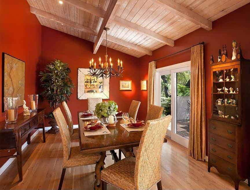 The wooden shed ceiling of this medium-sized dining room has exposed wooden beams matching with the hardwood flooring. These are contrasted by the earthy red tone of the walls that complements the dining room cabinet, console table and dining table perfectly paired with rustic woven wicker Parson's chairs.